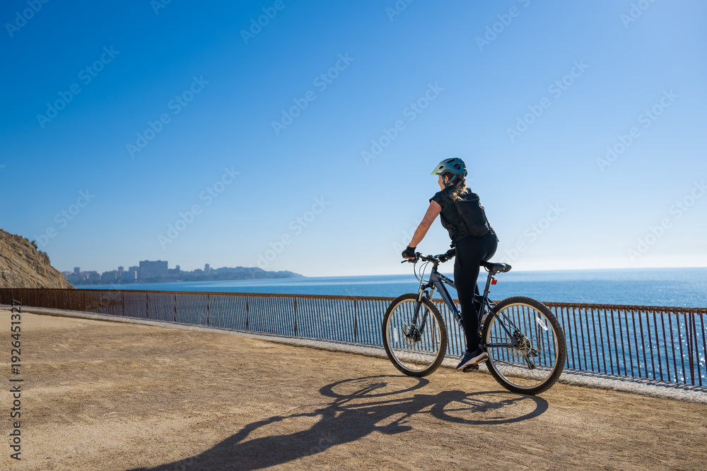 Obraz premium Rear view of female cyclist pedaling along Mediterranean waterfront trail with expansive sky and ocean backdrop, symbolizing exploration, independence and active outdoor lifestyle.