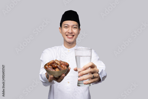 Young asian man presenting bowl of dates and glass of water for iftar, warm open expression, indoor studio scene, traditional