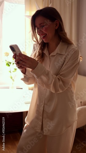 A vertical shot of a young woman in a silk shirt smiling and gesturing while having a video call or texting on her smartphone in a cozy, sunlit room.