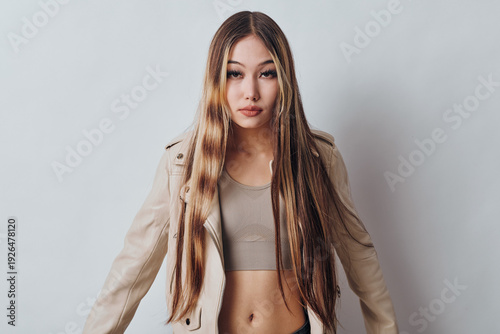 Young woman with long straight hair wearing beige cropped top and light jacket standing against plain white background looking at camera with neutral expression.