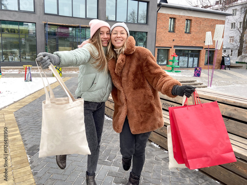 Two cheerful friends holding shopping bags outdoors in winter. Joyful mood, lifestyle, and shared happiness during city leisure and holiday activities.
