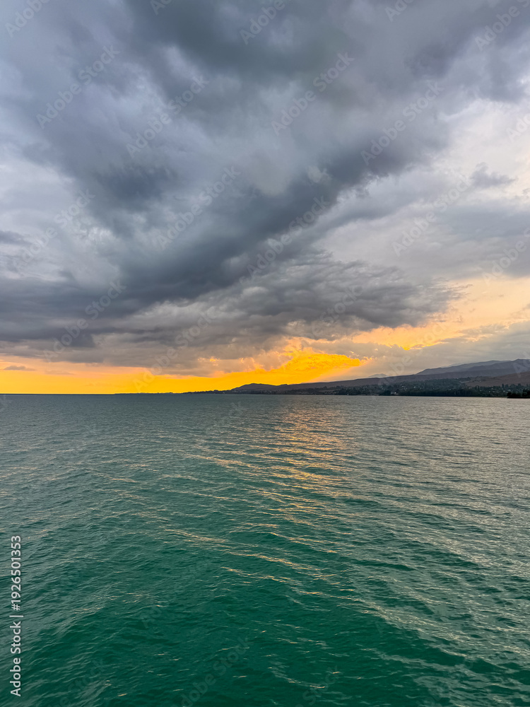 Fototapeta premium Sunset at Issyk Kul Lake in Kyrgyzstan. Bright sun breaking through clouds reflecting in clear turquoise water.