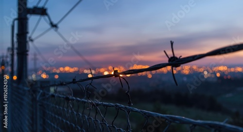 Power transmission cables at twilight behind a focused wire mesh fence and city blur