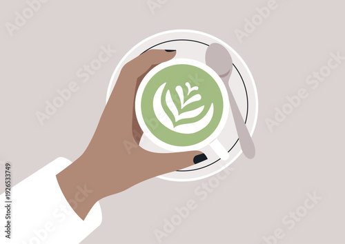 A hand gracefully holds a cup of matcha latte adorned with beautiful latte art, capturing a moment of warmth and comfort in a peaceful cafe setting.