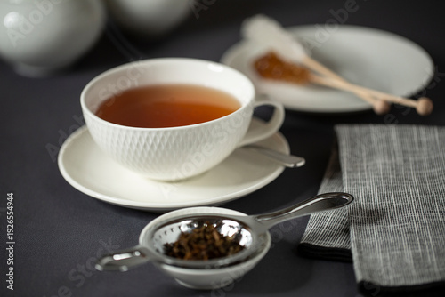Schwarzer Tee in einer weißen Porzellantasse auf einem Tisch. Die Tasse ist umgeben von einem teesieb, Kandiszucker am Stiel und einer Serviette.