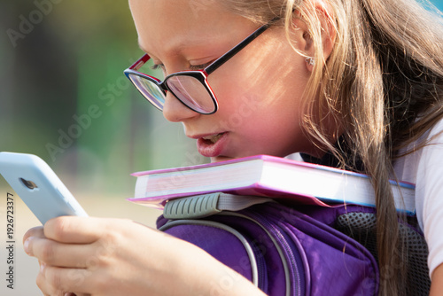 Cheerful portrait of a teenage schoolgirl with a phone and textbooks wearing reading glasses
