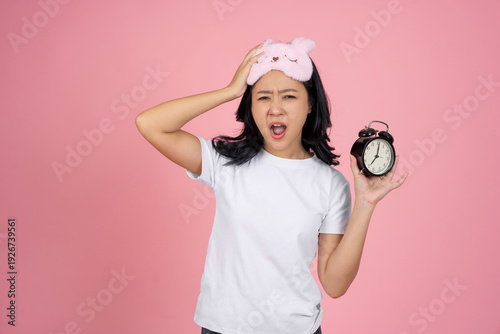 Sad Asian woman in pajamas and an eye mask is holding an alarm clock in the morning.