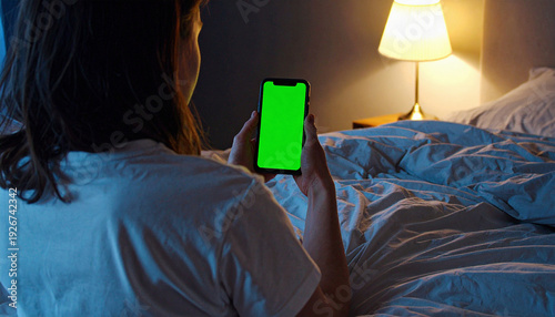 Mockup, green blank screen. Closeup of back view young white woman looking at her mobile phone and scrolling while lying in bed.
