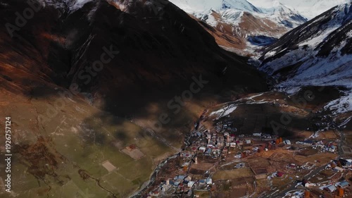 aerial view of Ushguli stone towers, ancient Svanetian village nestled on steep valley slope with scattered stone houses and defensive towers, late autumn light bathing terraced roofs and snow