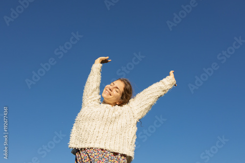 sweet, freedom-loving, happy young European woman enjoys the moment, raising her arms to the sun under a cloudless sky. love life, well-being, positive thinking, and dreams. A spring walk outdoors