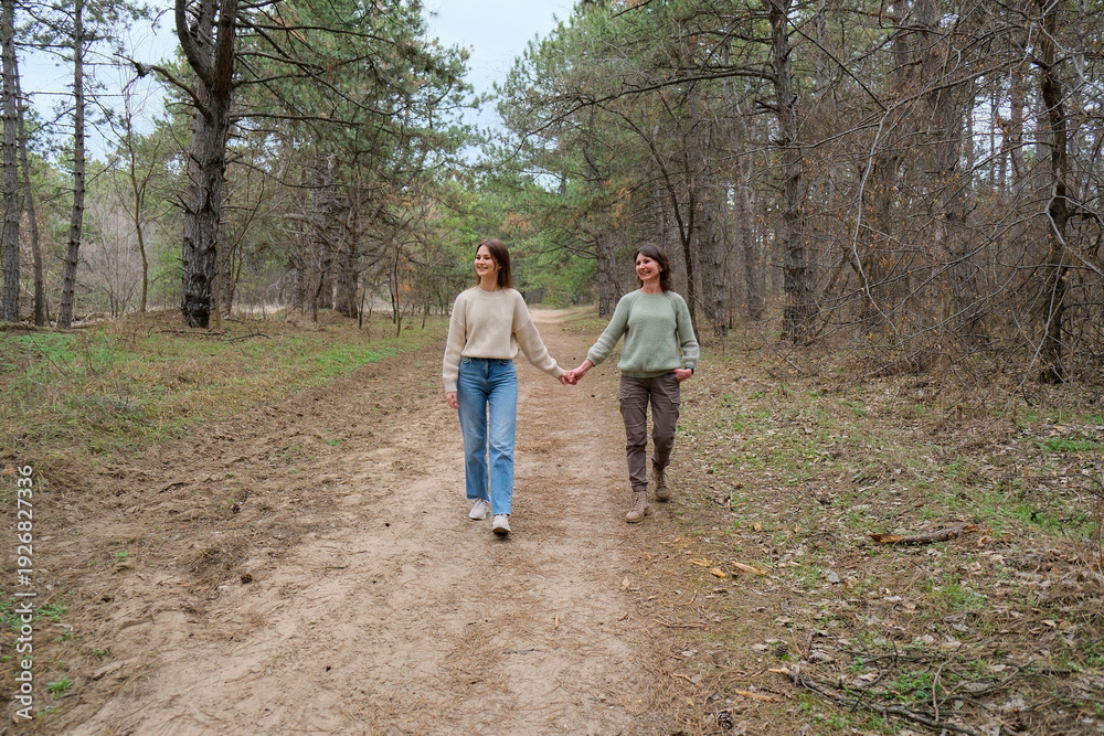 Fototapeta premium Mother and daughter are walking hand in hand along a dirt path in a natural park, enjoying their connection, family bond, and togetherness surrounded by trees and dry grass