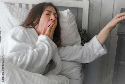 A sleepy middle-aged woman yawns while lying in bed under a white blanket. Early rise. Lack of sleep or disruption of the morning routine. Morning concept. Seasonal insomnia. Space for text. Banner