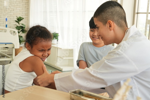 A pediatrician holds a syringe and administers a flu vaccine to a young African-American girl sitting with her father at the hospital.
