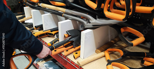 A person's hand selects one of the shovels on the store shelves. Concepts include home renovation, spring gardening preparation, construction work, DIY projects, and gardening.