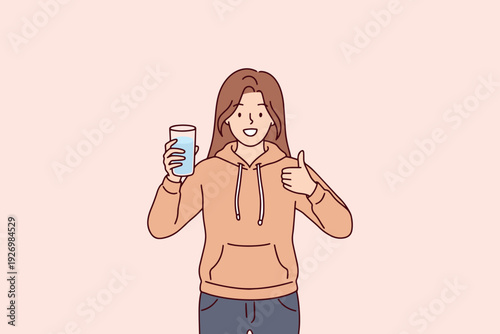 Happy young woman holds glass of water and gives thumbs up, representing body detox and healthy hydration. Concept of organic cleansing, health consciousness, and wellness routine for skin.
