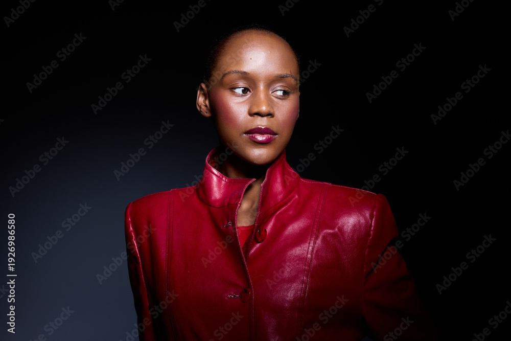 Obraz premium Portrait of a confident dark skinned African woman wearing a red leather jacket, looking sideways against a dark background. Concept of fashion, elegance, and empowerment.