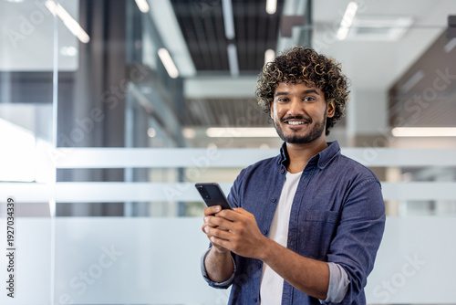 Portrait of a young Indian man standing in an office, holding a mobile phone and looking at the camera with a smile