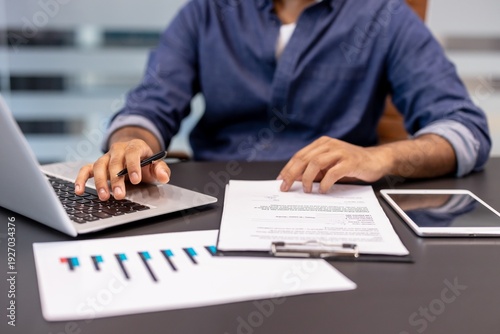 A close-up photo of the hands of a young man sitting at a desk in an office, working on a laptop and with documents