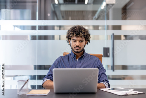 Serious and confused young Indian man sitting at a desk in the office and working on a laptop