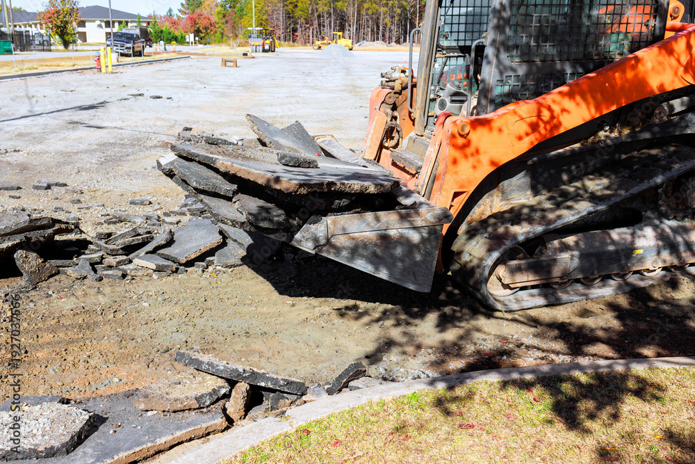 Fototapeta premium Skid steer machinery works to break up remove old asphalt at construction site in residential neighborhood.