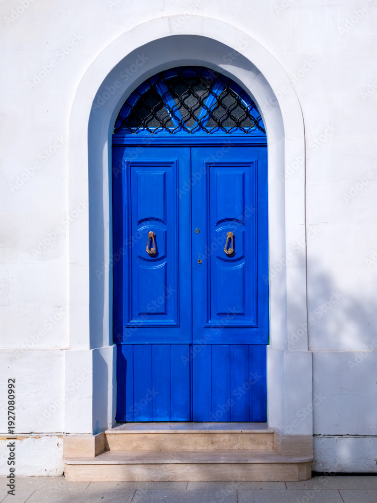 Fototapeta premium Blue double door with metal knockers under arch in white facade with stone steps.
