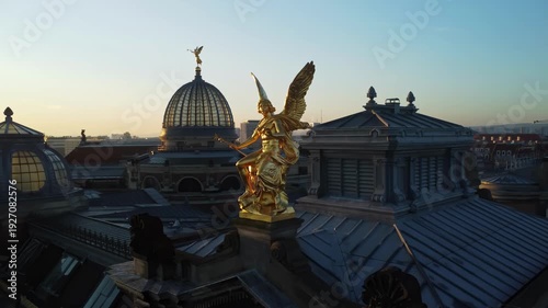 A circular aerial shoot of the statue of god Phantasos on the western corner risalit of the Dresden Art Academy  in the Lipsiusbau building under sunshine at dawn with the Frauenkirche, Dresden's Old 