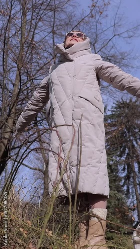 Vertical video. Relaxed elderly woman in long jacket with spread arms looks at sun standing on parapet in spring park. Happy senior lady enjoys nice day at winter end in city. Active retirement