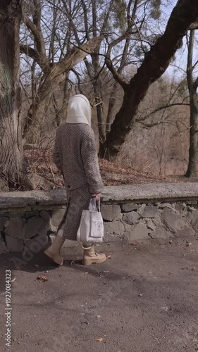 Vertical video. Senior woman in elegant coat and woolen scarf walks to rest on stone parapet in spring park. Healthy retired lady spends day in sunny urban garden. Activities at retirement time