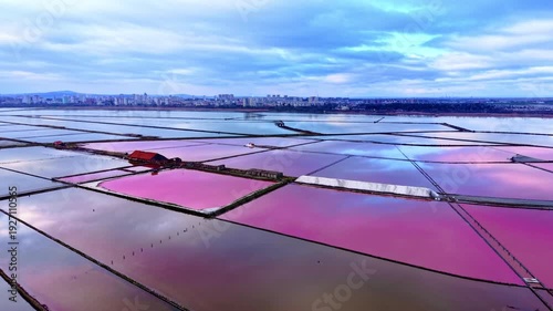 Geometric salt pans with vibrant pink water under a cloudy sky, city skyline in the distance. Aerial view of pink salt evaporation ponds near city coast.