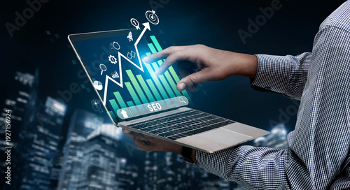 Fotografie Hands Holding Laptop with SEO Graph and Data Visualization Against City Skyline at Night for Digital Marketing Concepts Quark