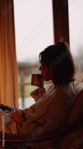 A woman is sitting in a chair with a cup of tea in her hand. She is looking out the window, possibly lost in thought. Concept of relaxation and contemplation