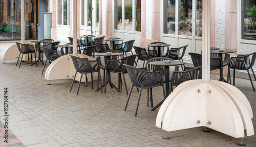 Empty outdoor restaurant terrace with black modern chairs, round tables, and large white umbrella bases on a paved street. Minimalist exterior