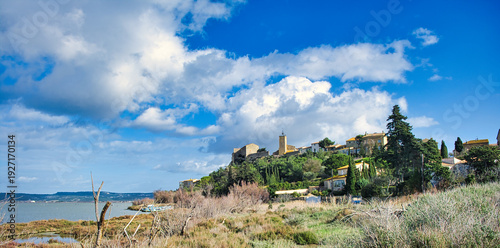 View of the picturesque Mediterranean village of Bages, department of Aude, France, perched on a hill on the shore of a lagoon under a cloudy sky.
