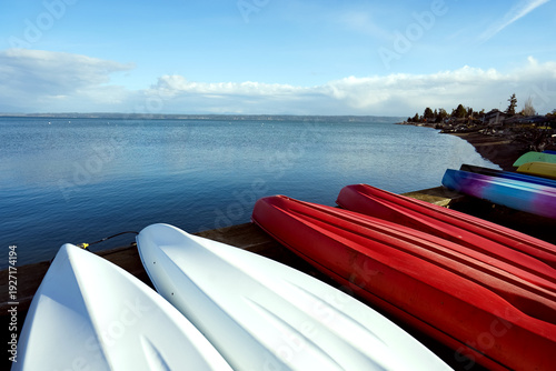 Colorful kayaks stored upside down on wooden pier by calm blue sea under soft cloudy sky with distant shoreline and coastal houses on background. Concept of summer recreation and waterfront lifestyle