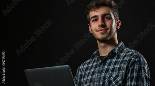 A young man in a plaid shirt working on a laptop against a dark background