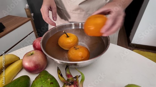 Hands of a woman in an apron standing in front of a table with fruit and washing mandarins