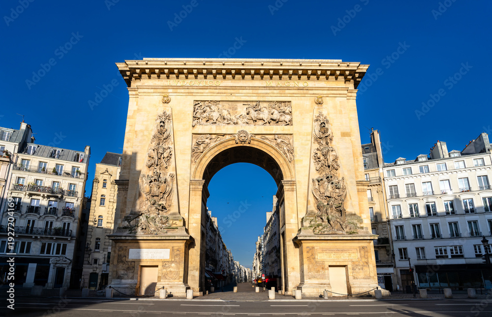 Obraz premium Scenic sunny view of the historic Porte Saint-Denis triumphal arch with intricate bas-relief carvings and classical Parisian buildings in Paris, France.
