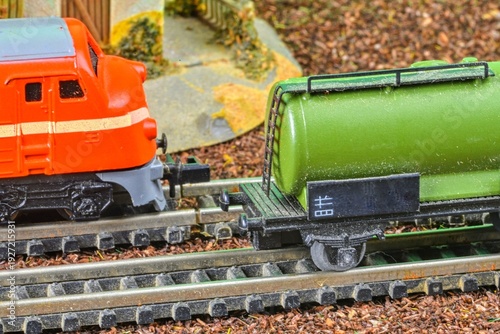 Close-up of a miniature red locomotive pulling a green tank car on model train tracks. Detailed view of a small-scale model train set featuring an orange engine coupled with a green liquid transport