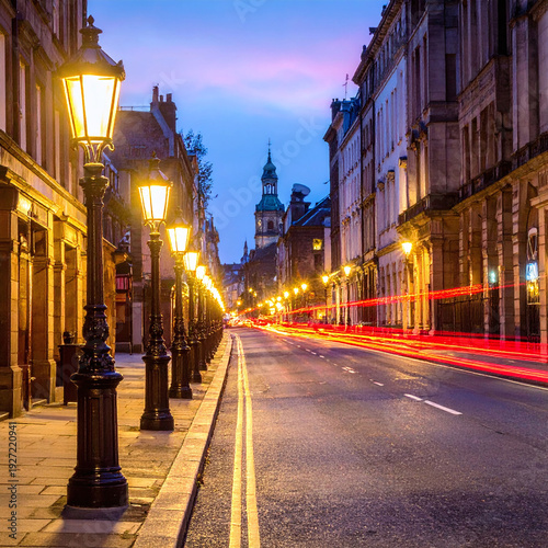 Enchanting evening view of a historic street with glowing vintage lamps and dynamic light trails. This atmospheric urban scene captures the timeless beauty of a European city at dusk.