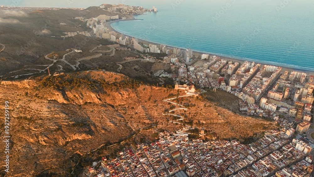 Fototapeta premium Aerial drone view of Cullera in Spain during golden hour, featuring a hilltop castle and winding path above the Mediterranean Sea. Detailed coastal cityscape with rooftops, shoreline and dramatic suns