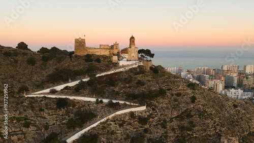 Aerial drone view of Cullera in Spain during golden hour, featuring a hilltop castle and winding path above the Mediterranean Sea. Detailed coastal cityscape with rooftops, shoreline and dramatic suns