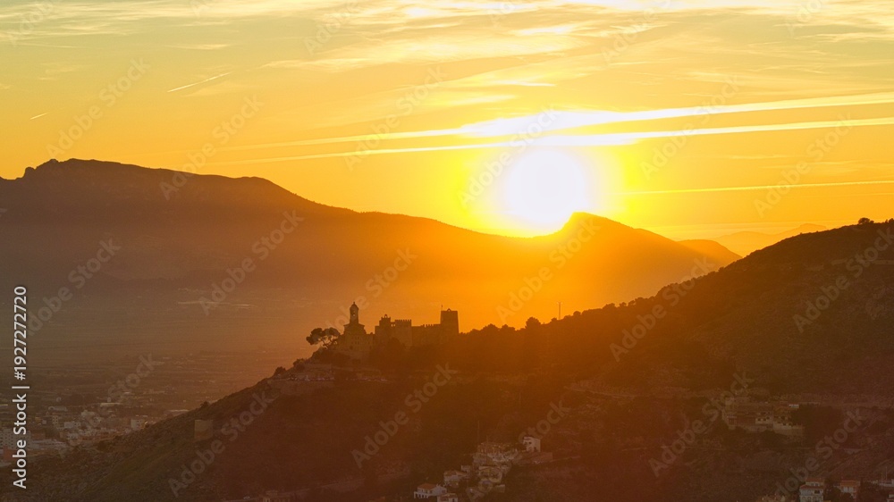 Obraz premium Cinematic aerial view of Cullera in Spain at sunset with castle and church silhouetted against golden light. Mediterranean coastline and mountain backdrop create a dramatic coastal cityscape.
