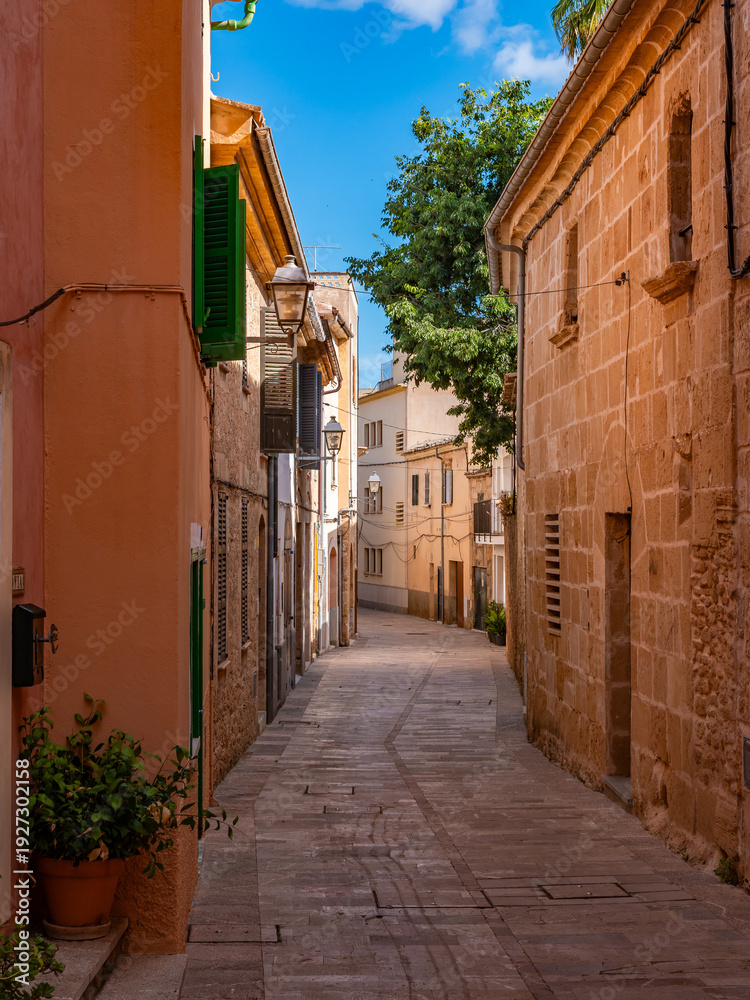 Fototapeta premium Narrow cobblestone alley winds through traditional Mediterranean buildings in Alcudia's old town, showcasing classic Mallorcan sandstone construction and peaceful residential charm.