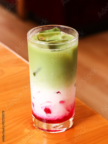 Three-layered iced matcha strawberry latte in a tall glass. Refreshing cold drink with red strawberry syrup, white milk, and green matcha tea layers on a wooden table.