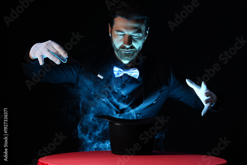 Fotografija Male magician with wand and hat making smoke at table on dark background