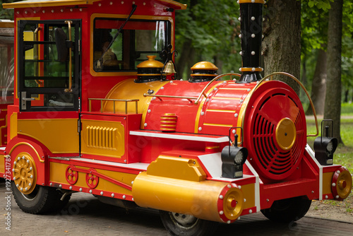 Colorful excursion train locomotive for children rides in city park with green trees background. Family entertainment, urban attraction, leisure activity and childhood recreation outdoors