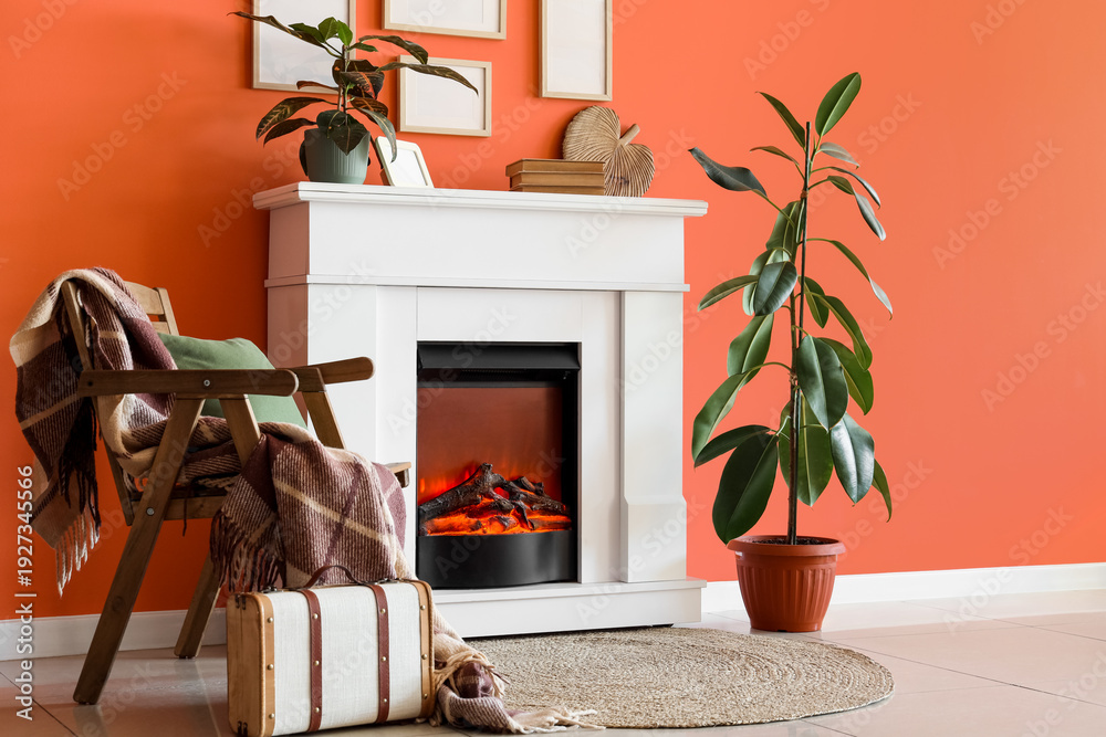 Obraz premium Fireplace with armchair, houseplants and photo frames near orange wall in living room