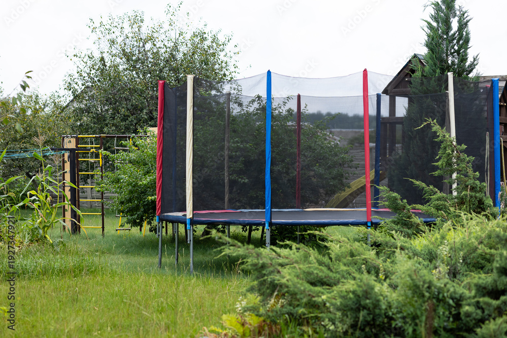 Fototapeta premium Backyard playground with trampoline and protective safety net in green garden near family house on sunny summer day. Safe active games, child protection, outdoor fitness, healthy lifestyle and fun