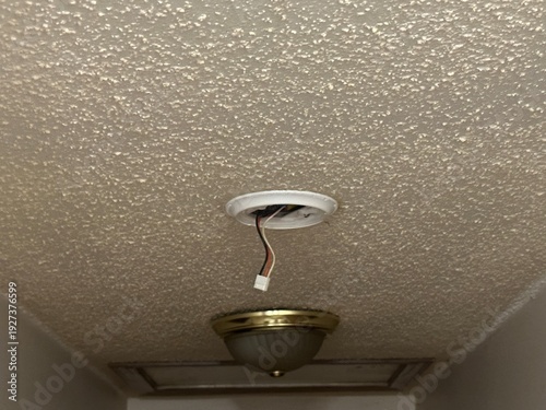 Close-up of an open recessed ceiling light fixture showing exposed electrical wiring and mounting hardware against a textured popcorn ceiling. Home maintenance, repair, and electrical installation