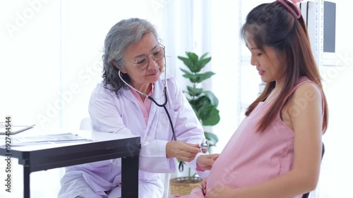 Elderly female doctor uses stethoscope to check pregnancy health of Asian woman. Health concept. Medical services.